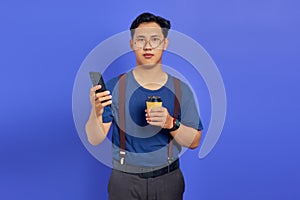 Sad handsome young man holding smartphone and cup of coffee on purple background