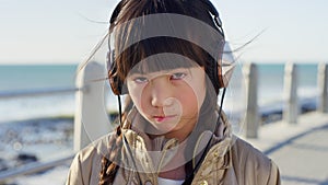 Sad, headphones and face of Asian girl by ocean with attitude, moody and angry expression listening to music. Childhood