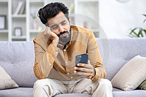 Sad hindu man sitting on sofa in bright room at home, holding phone in hands looking at screen, sad because of bad news