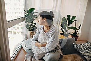 Sad hipster girl holding pillow, sitting at home during quarantine and looking at window. Stay home stay safe. Isolation at home