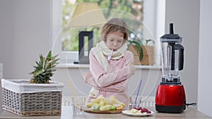 Sad ill Caucasian girl with thermometer in mouth standing at table with healthful fruits measuring body temperature