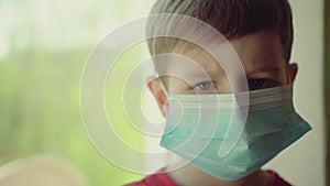 Sad illness child on home quarantine. Boy and his teddy bear both in protective medical masks sits on windowsill and looks out