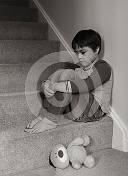 Sad kid sitting on staircase with teddy bear lying down on carpeted in house, Preschool boy looking down with upset face not happy