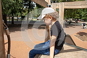 Sad little boy sits on the playground in the park. There is no one to play with. Children`s grievances. The photo