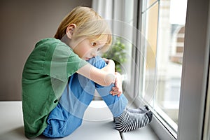 Sad little boy is sitting near window and watching street. Lonely at home. No friends, no siblings. One baby in family. Bad photo