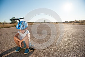 Sad little caucasian boy in helmet sitting on his scooter on asphalt empty road during summer hot sunset in California with copy