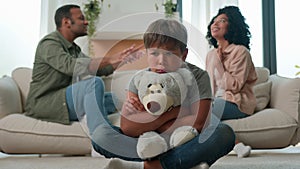 Sad little child boy hug teddy toy while mom dad fighting screaming arguing upset kid son suffer feel lonely parents