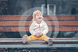 A sad little child is sitting on a bench on the playground alone. Rainy weather. The concept of lost childrens