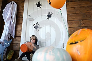 Sad little child in wizard costume with trick or treat candy basket sitting near door of house, decorated for Halloween.