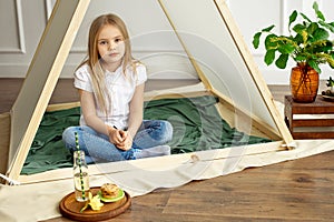 Sad little girl with blond hair in a white shirt and blue jeans is sitting in a tent in the children`s room.