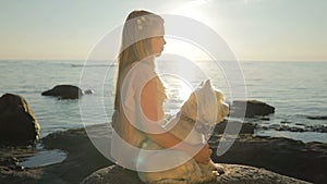 A sad little girl with a dog is sitting on the sea coast on the stones and watching the sunset.