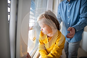 Sad little girl with Down syndrome looking throug window with her father at background.