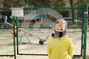 Sad little girl in face mask in yellow jacket standing near closed playground outdoor. Kids play area locked because of