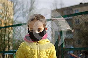 Sad little girl in face mask in yellow jacket standing near closed playground outdoor. Kids play area locked because of