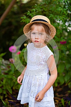 Sad little girl in hat, white dress among greenery