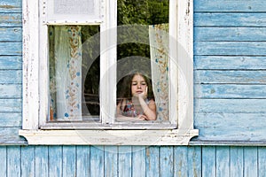 Sad little girl looking out the country house window leaning her face on her hand