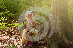 Sad little girl sitting on a stump inside the forest. Summer sunny day