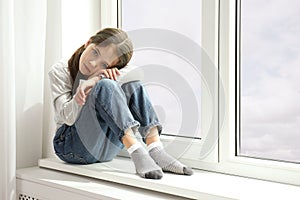 Sad little girl sitting on window sill, space for text