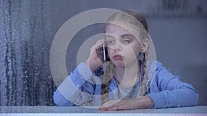 Sad little girl talking phone on rainy day, staying home alone, missing mother