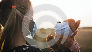 Sad little girl and teddy bear with surgical mask at sunset. A child holding her teddy bear during quarantine. Covid-19