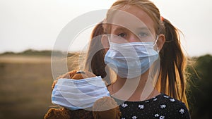 Sad little girl and teddy bear with surgical mask at sunset. A child holding her teddy bear during quarantine. Covid-19