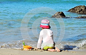 Sad little girl in a white shirt sits on the shore lonely plays with toys on the beach looks at the waves of the sea