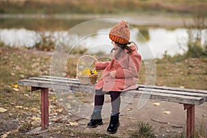 Sad little preschooler girl in a brown coat and orange hat sits on a wooden bench near the lake, looks at the basket to the left.