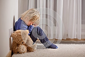 Sad little toddler child, blond boy, sitting in corner with teddy punished for mischief