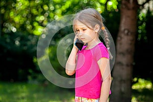 Sad lone little school age girl, child talking on the phone, smartphone outside. Unhappy, downhearted kid alone, phone call sad photo