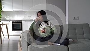 Sad lonely Asian woman sitting alone on sofa with cup of tea thinking of problems in personal life