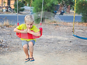 Sad lonely boy sitting on swing looking down on ground, waiting for friends or while parents are busy. Summer, childhood