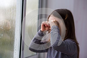 Sad lonely child girl with brown hair cries near the window. raindrops on glass. soft focus