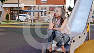 A sad lonely girl is sitting against the background of cheerful children playing on the playground.