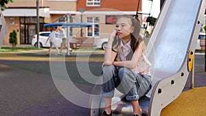 A sad lonely girl is sitting on a children's slide against the background of children having fun on the playground.