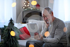 Sad and lonely senior man with tablet sitting at the table indoors at Christmas, solitude concept.