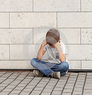Sad, lonely, unhappy, disappointed child sitting alone on the ground. Boy holding his head, look down. Outdoor