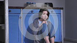 Sad lonely young Asian girl sitting on the floor in kitchen, holding her knees with arms, domestic violence concept 50