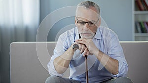 Sad male pensioner sitting on sofa, holding walking frame and looking down
