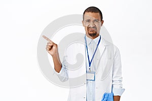 Sad male physician, doctor pointing left, grimacing and looking displeased, standing over white background