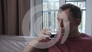 Sad man drinking alcohol while sitting on sofa, window background