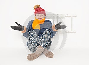 Sad man feeling cold in hat and blue sweater sitting close to radiator on white background