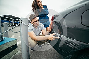 Sad man looking on car scratch, woman stand behind him