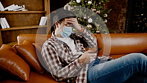 Sad man in mask surfing Internet alone at home on couch at Christmas time. Quarantine during New Year holidays ruined mood and