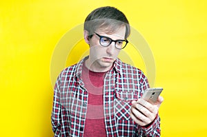 Sad man in red checkered shirt and glasses looking at smartphone on yellow background