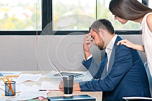A sad man sits at the desk and clings to his head, an employee stands beside him and comforts him.