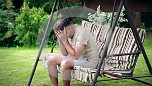 Sad man sits on a garden swing outdoors clutching his head and suffers for some reason