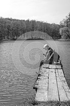 A sad man is sitting alone on pier by the lake. Forest black and white. hood on his head. backpack. in hands hold phone. Emotions