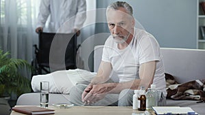 Sad man sitting on couch and looking into camera, male nurse bringing wheelchair