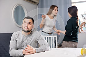 Sad man sitting at table at home while two women quarreling