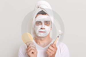 Sad man wearing white t shirt and towel on head posing isolated over gray background, holding sponge and massage roller in hands,